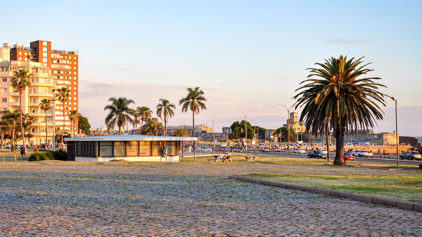 Rambla de Montevideo al atardecer, con palmeras, un pabellón bajo y edificios de fondo.