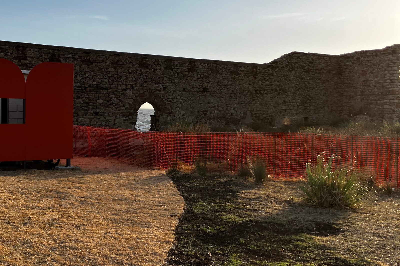Muro de piedra patrimonial del Espacio Mauá, con un arco que deja ver el agua, un pabellón rojo y el vallado del predio.